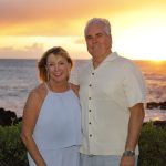 A smiling couple stands together outdoors in front of the ocean at sunset with greenery in the foreground.