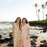 Two women in light dresses stand barefoot on a sandy beach with black rocks, ocean waves, and palm trees in the background.