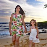 Woman and young girl holding hands and walking on a sandy beach with the ocean and colorful sky in the background.