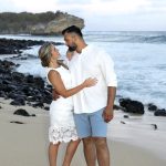 A couple stands barefoot on a sandy beach near black rocks, facing each other and smiling, with waves in the background.