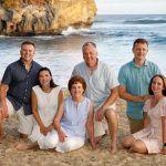 A group of six adults pose together on a sandy beach with a rocky cliff and ocean in the background.