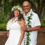 A couple wearing leis and white clothing stands outdoors, smiling at the camera with trees in the background.