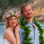 A couple poses by the ocean; the woman wears a flower crown and the man has a leafy lei around his neck.