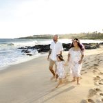 A family of three walks along a sandy beach near the shoreline, leaving footprints in the sand on a sunny day.