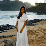 Woman in a white dress stands barefoot on a sandy beach with waves, rocks, and mountains in the background.