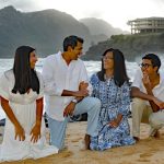 Four people kneel and smile together on a sandy beach with mountains and a building in the background.