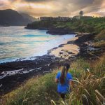 A woman in a blue shirt stands on grassy cliffs overlooking a sandy beach, ocean waves, and distant houses at sunset.
