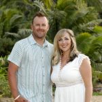 A man and woman stand together outdoors in front of tropical plants, both smiling at the camera.