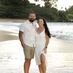 A man and woman stand barefoot on a sandy beach with waves and greenery in the background.
