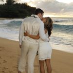 A couple dressed in white wedding attire kisses on a sandy beach at sunset, facing the ocean.